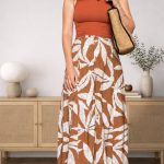 petite woman in an orange ribbed tank top and a rust-and-cream leaf-print maxi skirt, a raffia shoulder bag over one arm, standing in front of a light rattan sideboard.