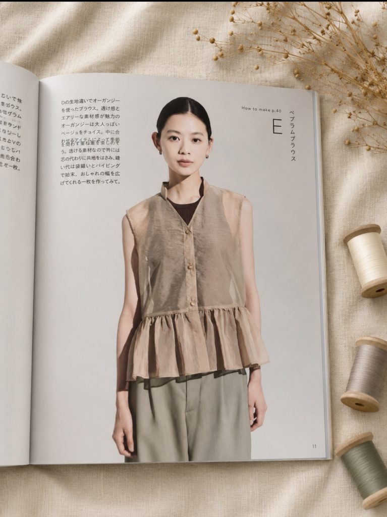a model in a sheer beige organza sleeveless peplum blouse over grey trousers.