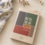 the paperback 'a dictionary of color combinations' (配色事典) lying on natural linen next to a sprig of dried flax, its cover divided into grey, green, red and brown colour blocks.