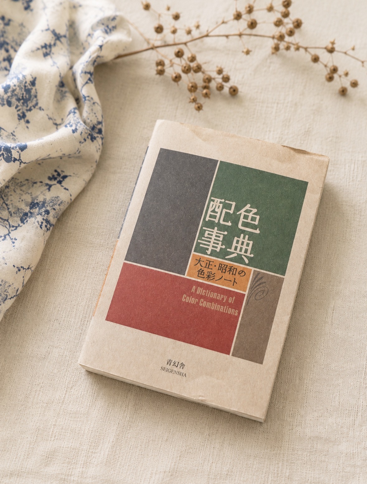 the paperback 'a dictionary of color combinations' (配色事典) lying on natural linen next to a sprig of dried flax, its cover divided into grey, green, red and brown colour blocks.