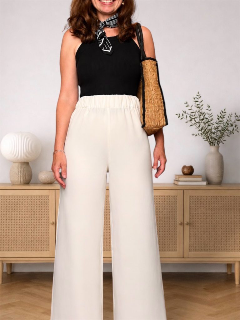 petite woman in the same black top and cream wide-leg trousers, holding a raffia "bella isola" tote in both hands, in front of a rattan sideboard.